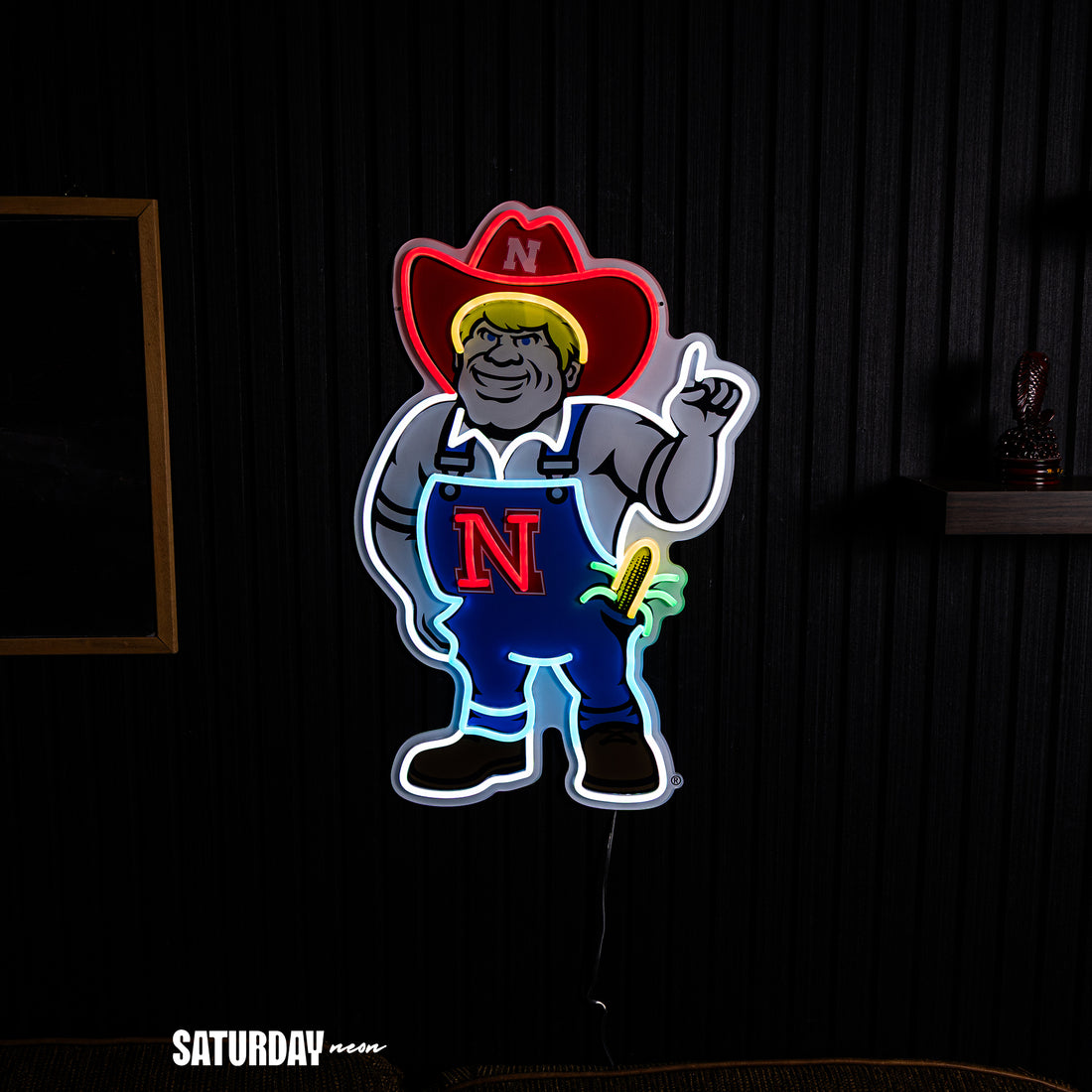 A neon-style wall sign featuring a Nebraska Cornhuskers mascot, Herbie the Husker. He's wearing a hat with a white 'N' and blue overalls with a large red 'N' on the chest, set against a dark paneled wall.